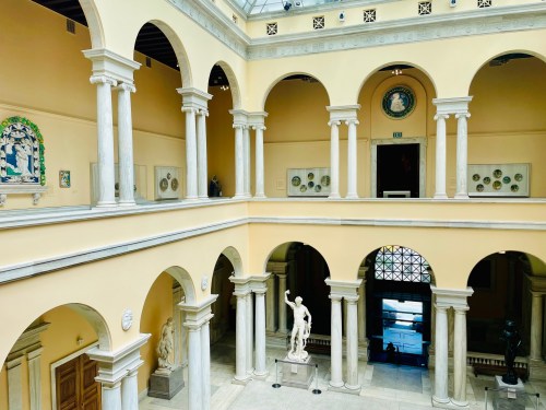 Interior view of a museum featuring tall columns, a statue, and artwork displayed on the walls. Natural light from above.