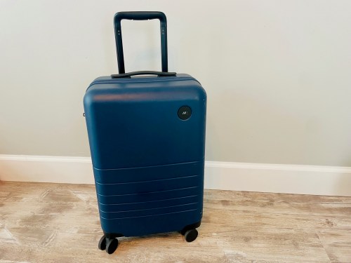 A blue hard-shell suitcase with a retractable handle and wheels, standing against a light-colored wall.