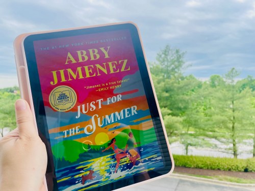 A hand holds a tablet displaying the book "Just for the Summer" by Abby Jimenez against a cloudy sky and greenery.