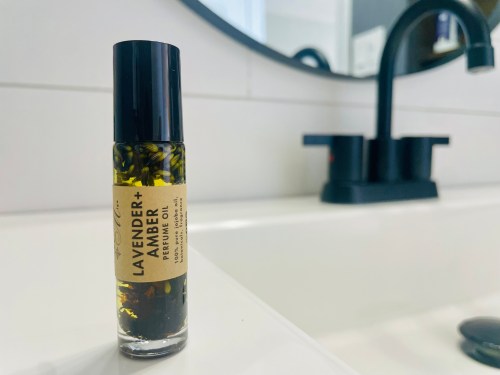 A bottle of lavender and amber perfume oil on a bathroom counter, with a black faucet in the background.