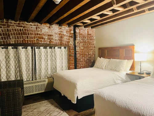 Cozy bedroom with two beds, exposed brick walls, wooden beams, and a lamp beside a window with patterned curtains.
