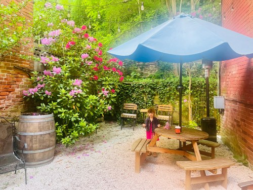A child stands by a wooden table under an umbrella in a garden with blooming flowers and brick walls.