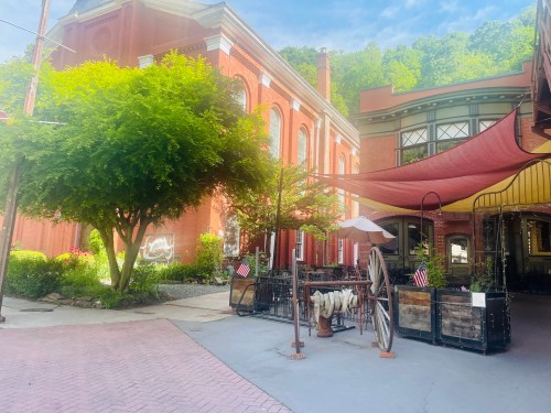 A charming outdoor area with red brick buildings, greenery, and a shaded seating space with American flags.