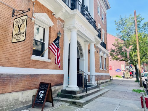 Exterior of a brick building with an American flag, featuring a sign for "The Spa" and a chalkboard menu outside.