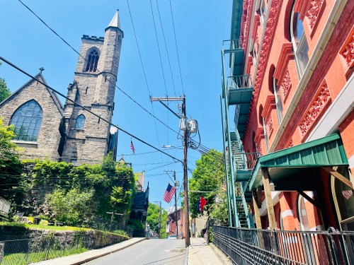 A historic church towers over a street lined with red brick buildings and flags under a clear blue sky.