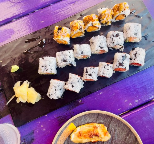 A platter of assorted sushi rolls on a black serving board, accompanied by pickled ginger and a side of dumplings.