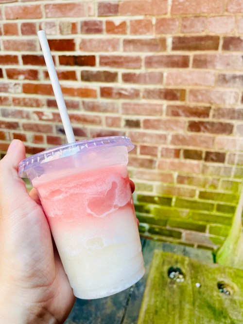 A hand holding a layered frozen drink in a clear cup, featuring pink and white swirls, against a brick wall background.