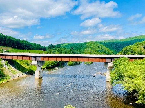 A bridge spans a river surrounded by lush green hills under a blue sky with scattered clouds.