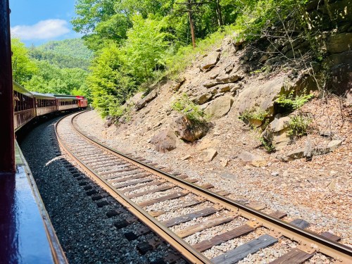 A scenic view of a train track curving through lush greenery and rocky terrain under a bright blue sky.