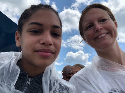 Two people wearing clear ponchos smile for a selfie against a backdrop of clouds.