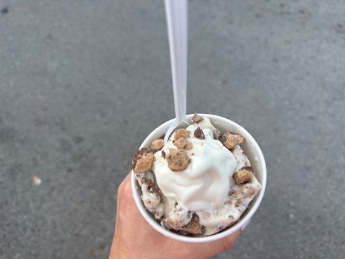 A hand holding a cup of ice cream topped with whipped cream and chocolate pieces against a gray background.