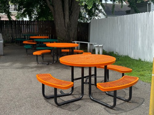 Colorful outdoor picnic tables with orange and green seats in a shaded area surrounded by a wooden fence.