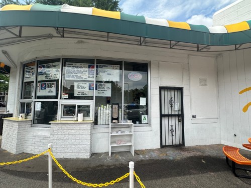A small food stand with a menu displayed in windows, featuring a green and yellow awning and outdoor seating.