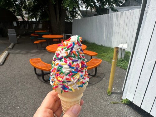 A hand holds a colorful ice cream cone topped with sprinkles, with outdoor seating in the background.
