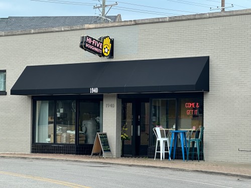 Exterior of Hi-Five Doughnuts shop with a black awning, bright sign reading "COME & GET IT," and outdoor seating.