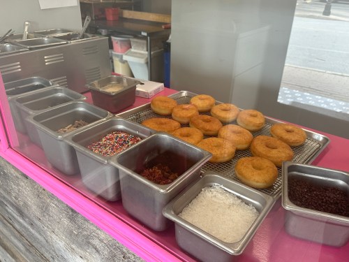 A display of freshly made donuts and various toppings in metal containers, viewed through a glass window.