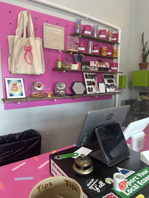 A colorful shop counter with a pink wall, displaying merchandise, a cash register, and a tip jar.