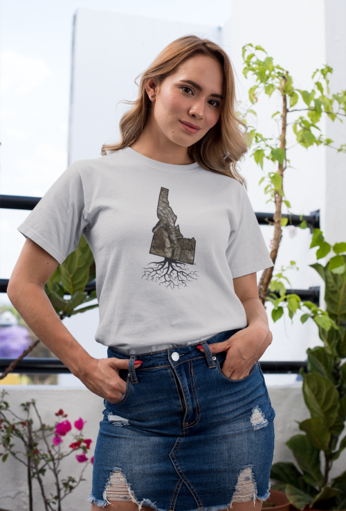 A woman wearing a gray t-shirt with a graphic of Idaho and roots, standing in a bright outdoor setting with plants.