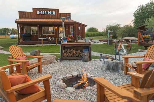A cozy outdoor scene featuring a fire pit surrounded by wooden chairs, with a rustic saloon in the background.