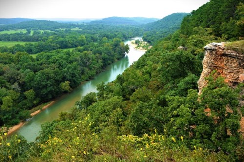 A scenic view of a winding river surrounded by lush green trees and hills under a cloudy sky.