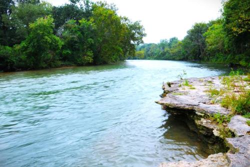 A serene river flows through a lush green landscape, with rocky banks and trees lining the water's edge.