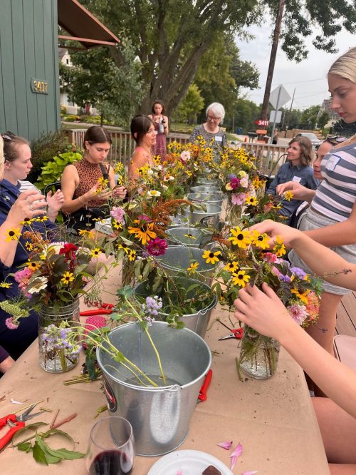 A group of people arranging flowers in jars on a table outdoors, surrounded by greenery and a relaxed atmosphere.