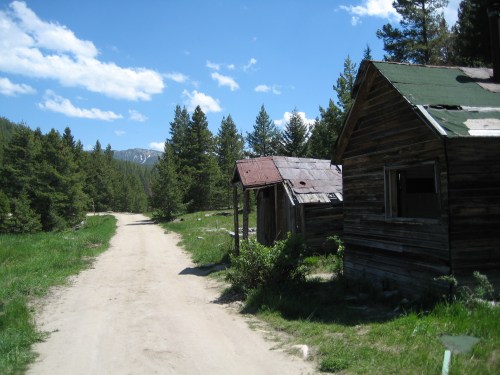 A dirt road leads past two rustic wooden cabins surrounded by tall trees and a clear blue sky.