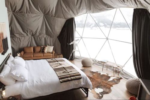 Cozy geodesic dome interior with a bed, sofa, and large window showcasing a snowy landscape. Warm, modern decor.
