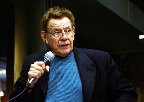 A man with glasses and a mustache speaks into a microphone, wearing a dark blazer over a blue sweater.
