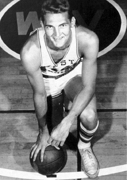 A young male basketball player kneels on the court, holding a basketball, smiling at the camera.