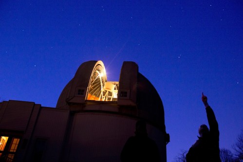 A silhouette of a person pointing at the night sky near a brightly lit observatory under a starry backdrop.