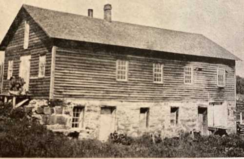 Historic two-story building with wooden siding and stone foundation, surrounded by grass and trees.