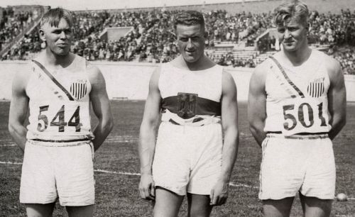 Three male athletes in vintage sports uniforms stand together on a field, with a crowd in the background.