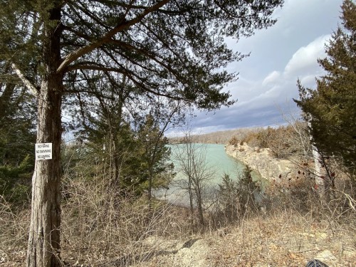 A scenic view of a river surrounded by trees, with a sign visible on a nearby tree. Cloudy sky in the background.