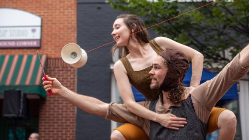 A performer balances on another's shoulders, skillfully juggling a diablo in a lively outdoor setting.