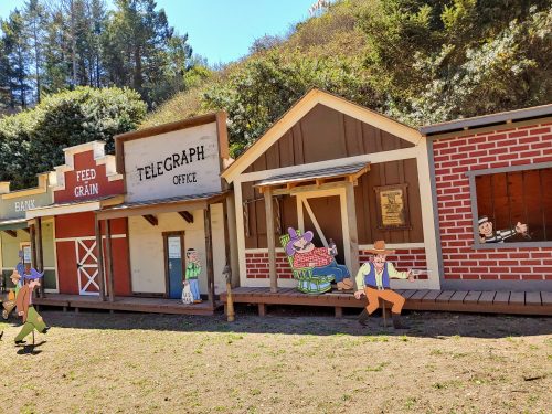 A colorful western-style town with cartoon characters, including a mouse and cowboys, in front of wooden buildings.