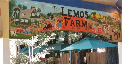 Colorful sign for "The Lemos Farm" featuring a farm scene with animals, a barn, and a train. Green umbrellas below.