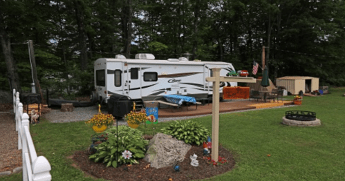 A parked RV in a green yard with flowers, a fire pit, and a small shed in a wooded area.
