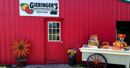 A red barn with a sign for Gieringer's Orchard, featuring a cart of fresh produce and colorful flowers outside.