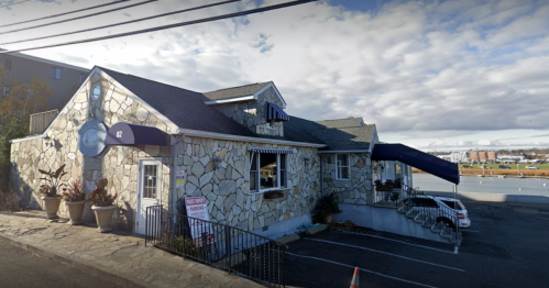 A stone-clad restaurant with a blue awning, surrounded by planters, and parked cars in the lot. Cloudy sky above.