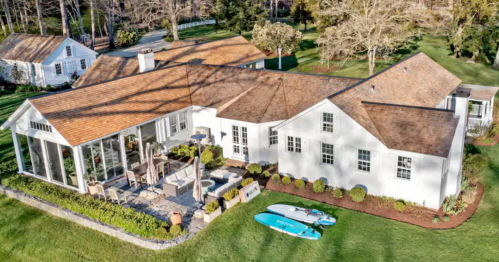 Aerial view of a modern white house with a patio, surrounded by greenery and two paddleboards on the lawn.