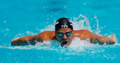 A swimmer in a black cap and goggles performs the butterfly stroke in a bright blue pool.