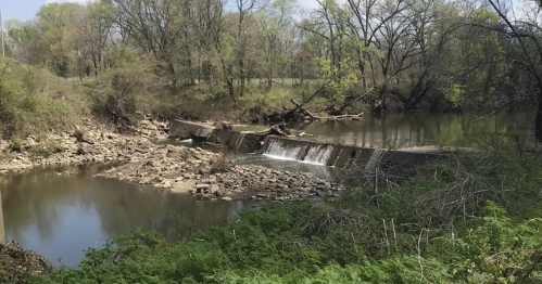 A serene river scene with a small dam, surrounded by trees and rocky banks, under a clear sky.