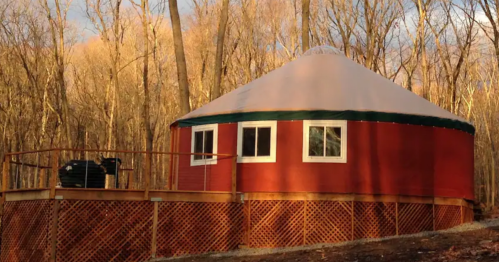 A red yurt with a white roof, surrounded by trees, on a wooden deck in a forested area.
