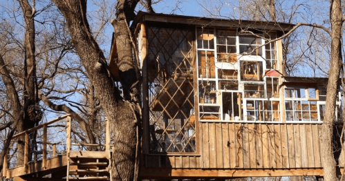 A unique treehouse made of wooden planks and various windows, nestled among bare trees against a clear blue sky.
