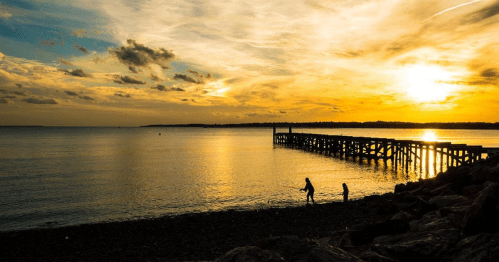 Silhouettes of two people walking along a rocky shore at sunset, with a pier extending into the calm water.