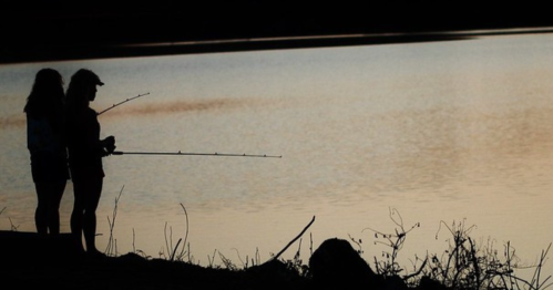 Two silhouetted figures stand by a calm lake at sunset, holding fishing rods and gazing at the water.