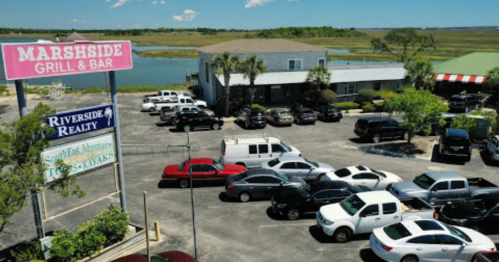 Aerial view of Marshside Grill & Bar with a parking lot filled with cars and a scenic landscape in the background.