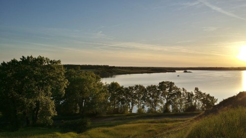 A serene landscape featuring a river at sunset, surrounded by lush trees and a clear sky.
