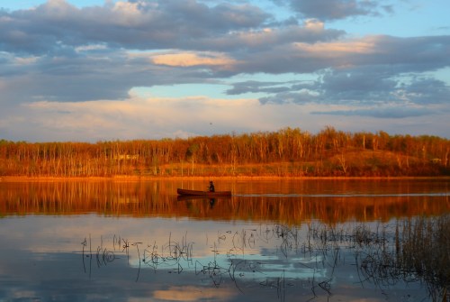 A lone person in a small boat glides across a calm lake, surrounded by trees and a colorful sunset sky.
